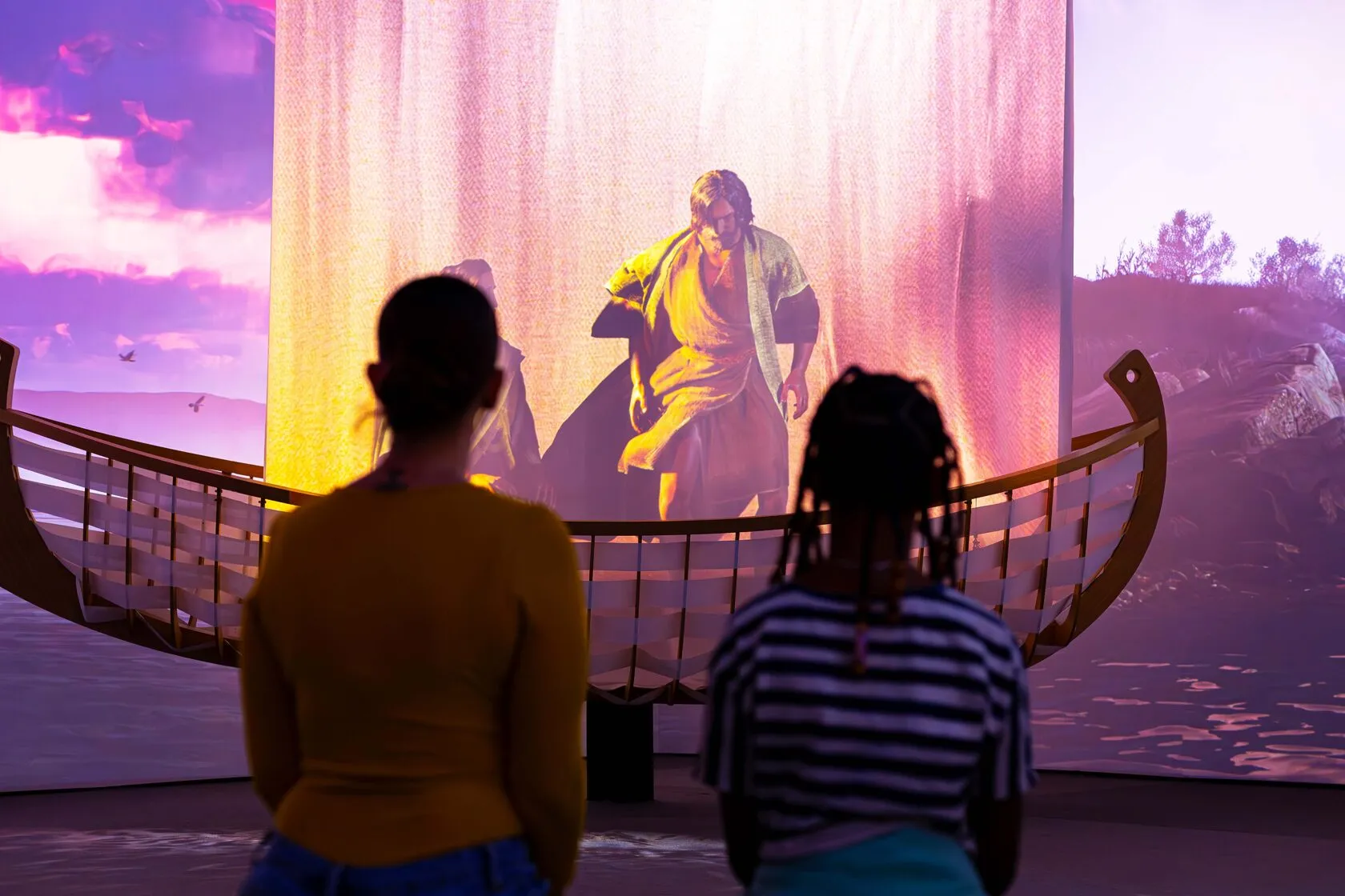 Two visitors watching a projected scene of a figure stepping into a boat at sunset.